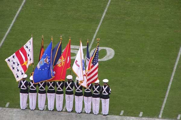 ROTC cadets lined up at the sideline of the 50-yard-line of a football field. They bear the flags of the united states, of the commonwealth of Virginia, and of the various armed services. (Credit: Bigstock)